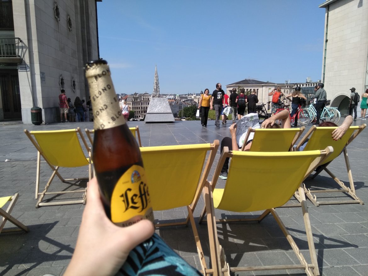 Woman's hand holding a beer up in front of yellow deckchairs on a hill overlooking Brussels city centre